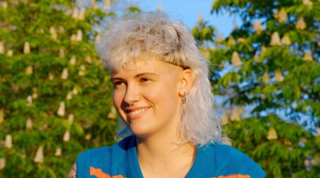 Una giovane donna con un mullet riccio biondo platino, seduta all'aperto in una giornata di sole.