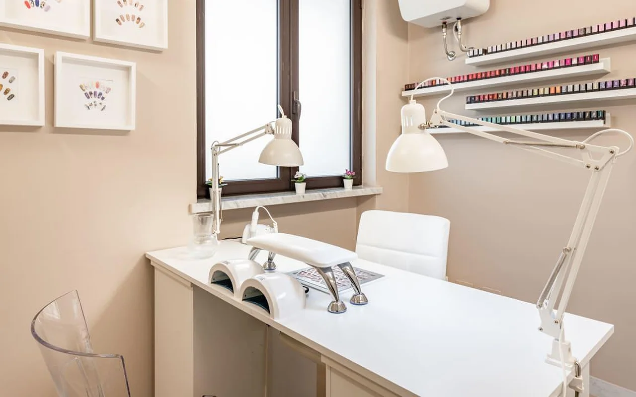 Una postazione dedicata alle unghie da AG The Beauty Room's, con una scrivania bianca e pulita, doppie lampade UV, luci da lavoro regolabili e mensole che espongono file di smalti colorati. Alle pareti dai toni caldi sono appesi disegni incorniciati di nail art.