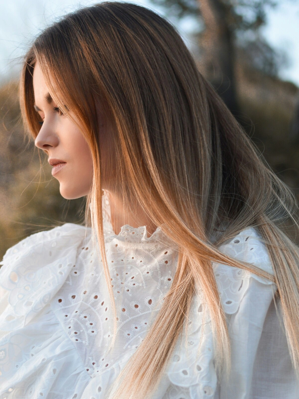 Veduta laterale di una donna all'aperto in un ambiente naturale, con il balayage su capelli lisci in cui il castano sfuma in tonalità bionde