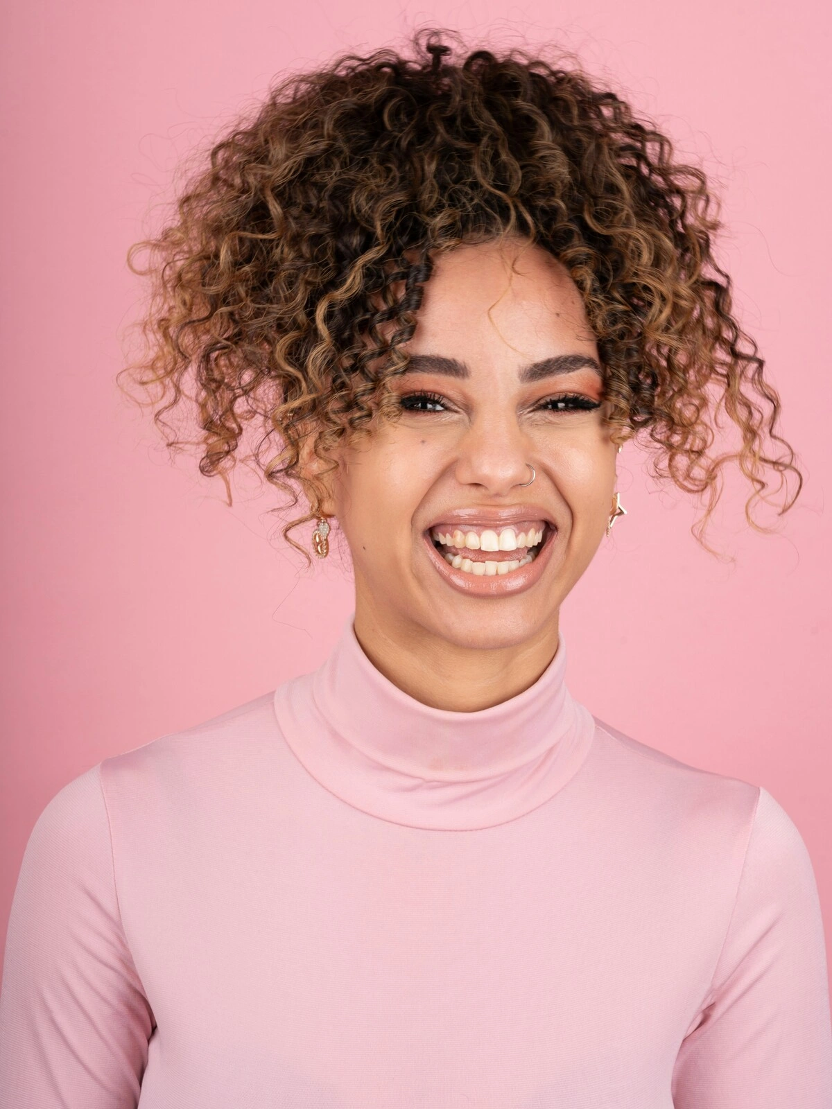Foto in studio di una donna sorridente con il balayage su capelli afro, vestita con un dolcevita rosa tenue