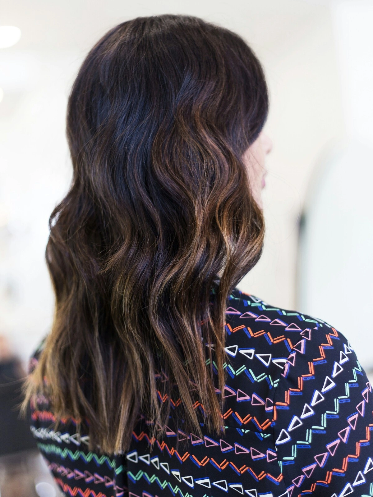 Veduta posteriore della testa di una donna con il balayage in onde morbide, in cui i capelli neri sfumano in tonalità castane calde