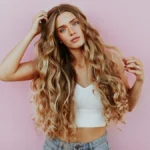 Foto in studio di una donna con capelli lunghissimi, mossi e baciati dal sole con il balayage, vestita con jeans azzurro chiaro e un top corto bianco a maglia