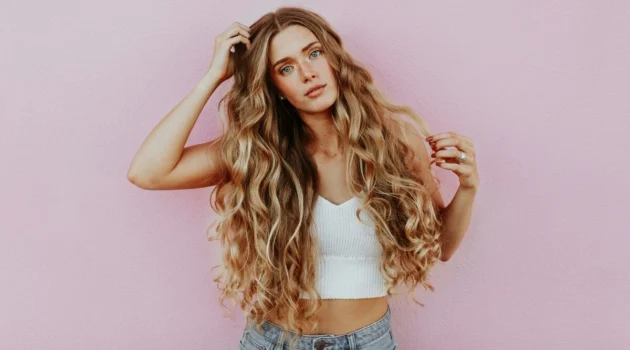 Foto in studio di una donna con capelli lunghissimi, mossi e baciati dal sole con il balayage, vestita con jeans azzurro chiaro e un top corto bianco a maglia