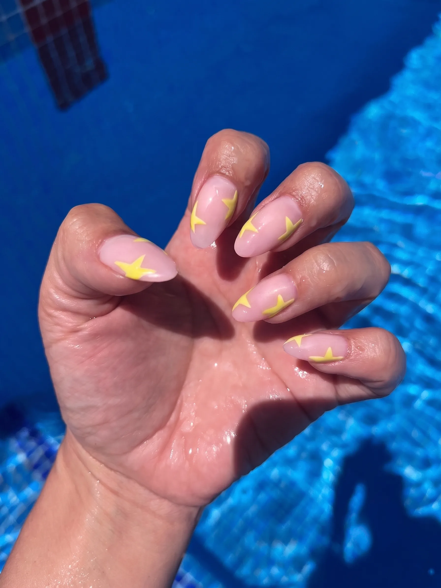 Unghie a mandorla in nude trasparente con stelle giallo burro, fotografate a bordo piscina.