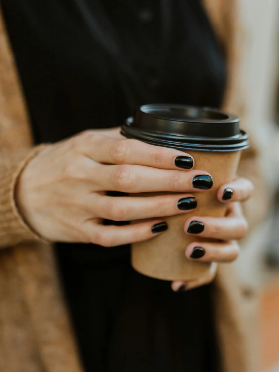 Primo piano di una donna con una manicure in gel nera e lucida che tiene un bicchiere di caffè da asporto