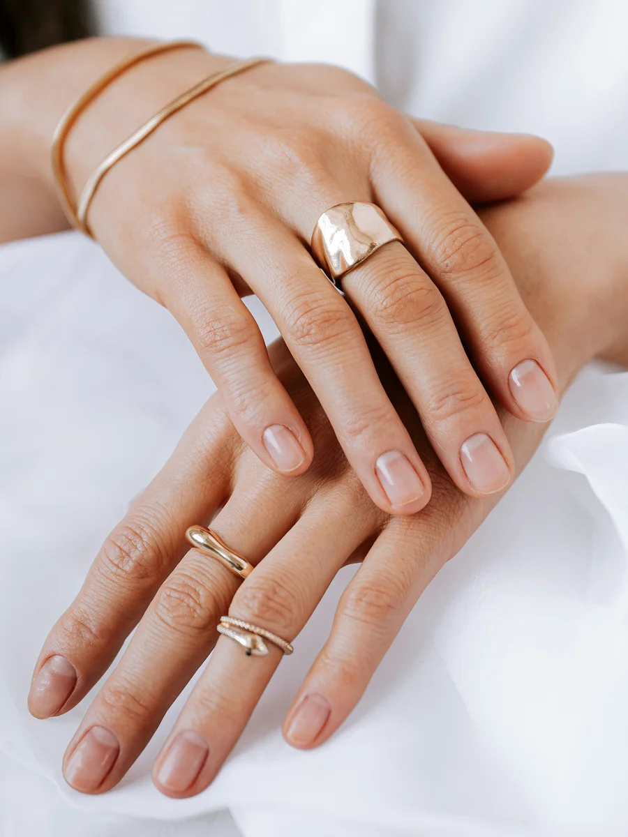 Primo piano di due mani appoggiate su un lenzuolo bianco che mostrano unghie naturali e sane, con anelli e bracciali color oro rosa