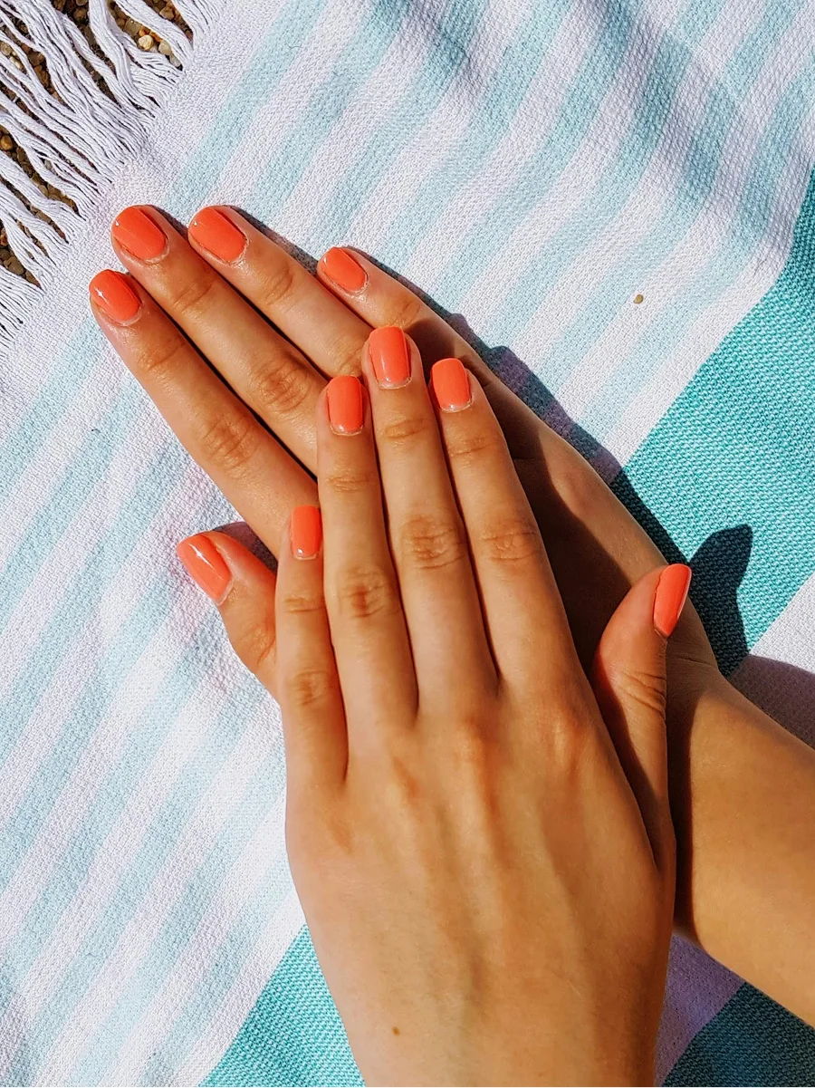 Vista dall’alto di due mani con una manicure in gel arancione lucida, appoggiate su un telo da mare a righe blu e bianche