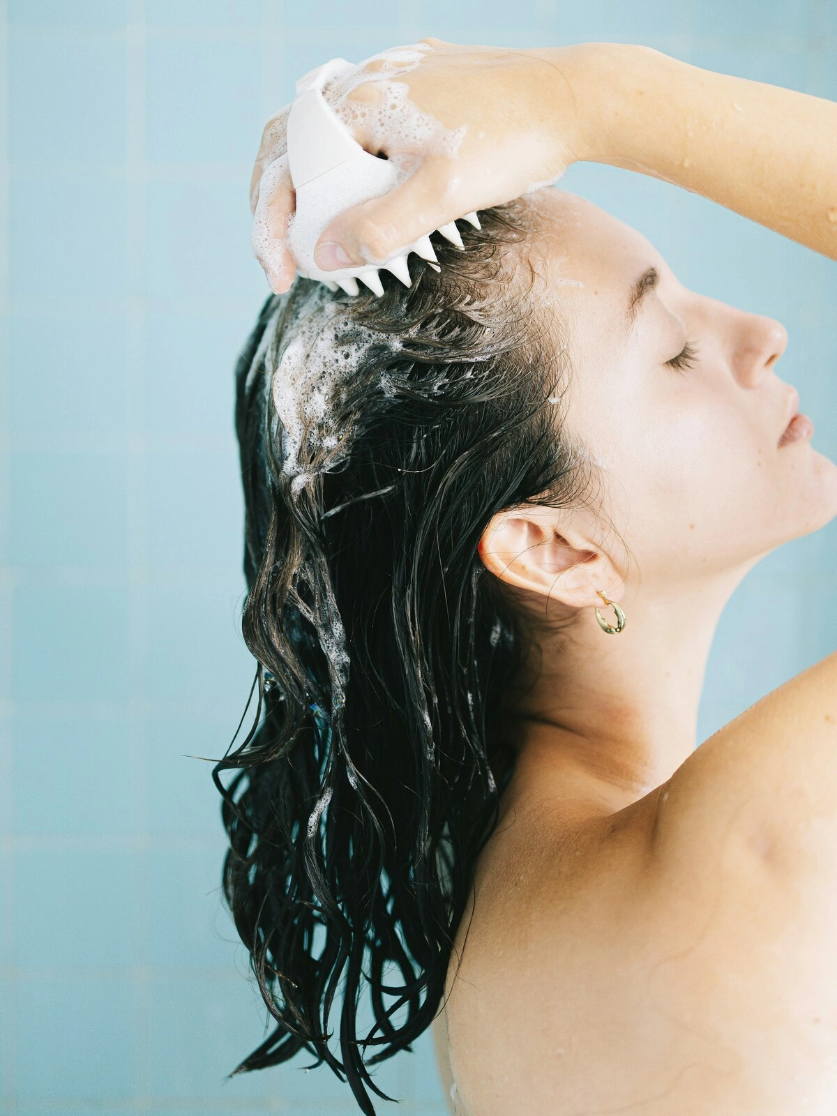 Veduta laterale di una donna con i capelli bagnati e insaponati che si massaggia il cuoio capelluto con una spazzola in silicone