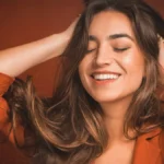 Primo piano di una donna che sorride con gli occhi chiusi mentre passa le mani tra i capelli castani, lunghi e morbidi