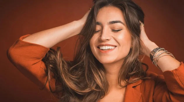Primo piano di una donna che sorride con gli occhi chiusi mentre passa le mani tra i capelli castani, lunghi e morbidi