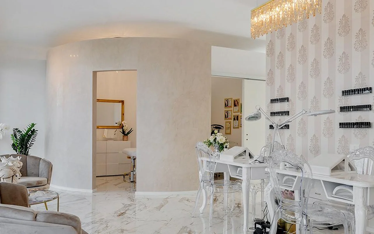 L'elegante interno di MC Beauty Center, con pavimento effetto marmo, pareti curve bianche e un lampadario dorato di cristallo. Sulla destra una postazione per le unghie con sedie trasparenti, mentre poltrone in velluto e una porta dalla cornice dorata conducono a una cabina trattamenti.