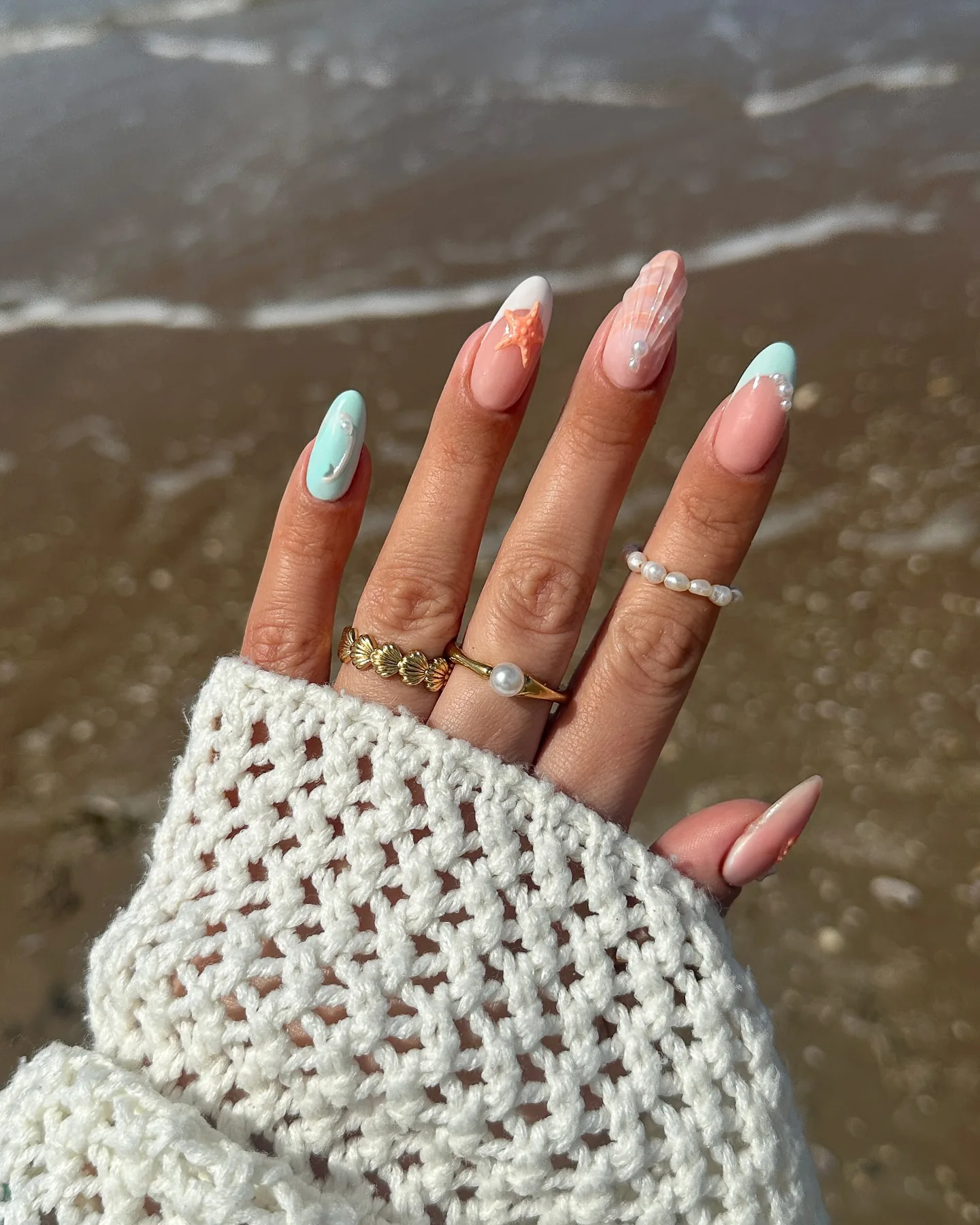 Unghie a mandorla con nail art sirena in tonalità pastello – una stella marina, accenti madreperlati e un bagliore iridescente – su sabbia bagnata.