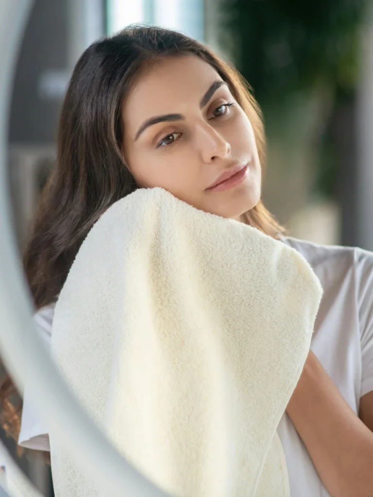 Una donna con la pelle fresca e luminosa e lunghi capelli scuri si asciuga delicatamente il viso con un asciugamano guardandosi allo specchio