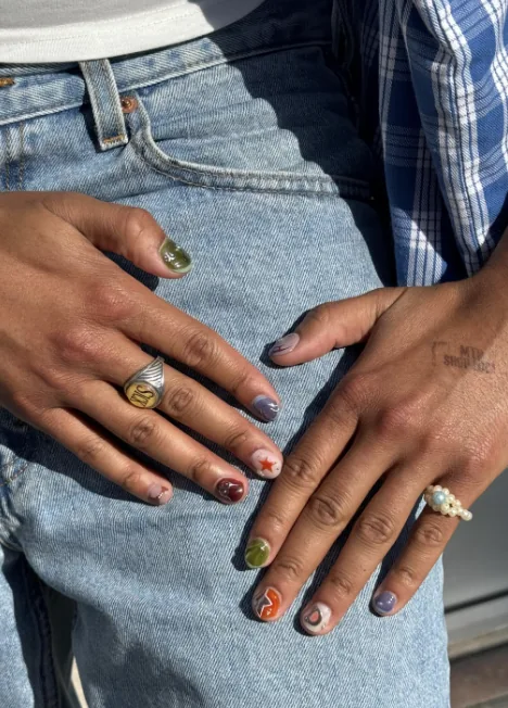 Manicure mismatched su unghie corte, ogni unghia di colore e disegno diversi, con una stella rossa e una voluta arancione, appoggiata su un jeans.