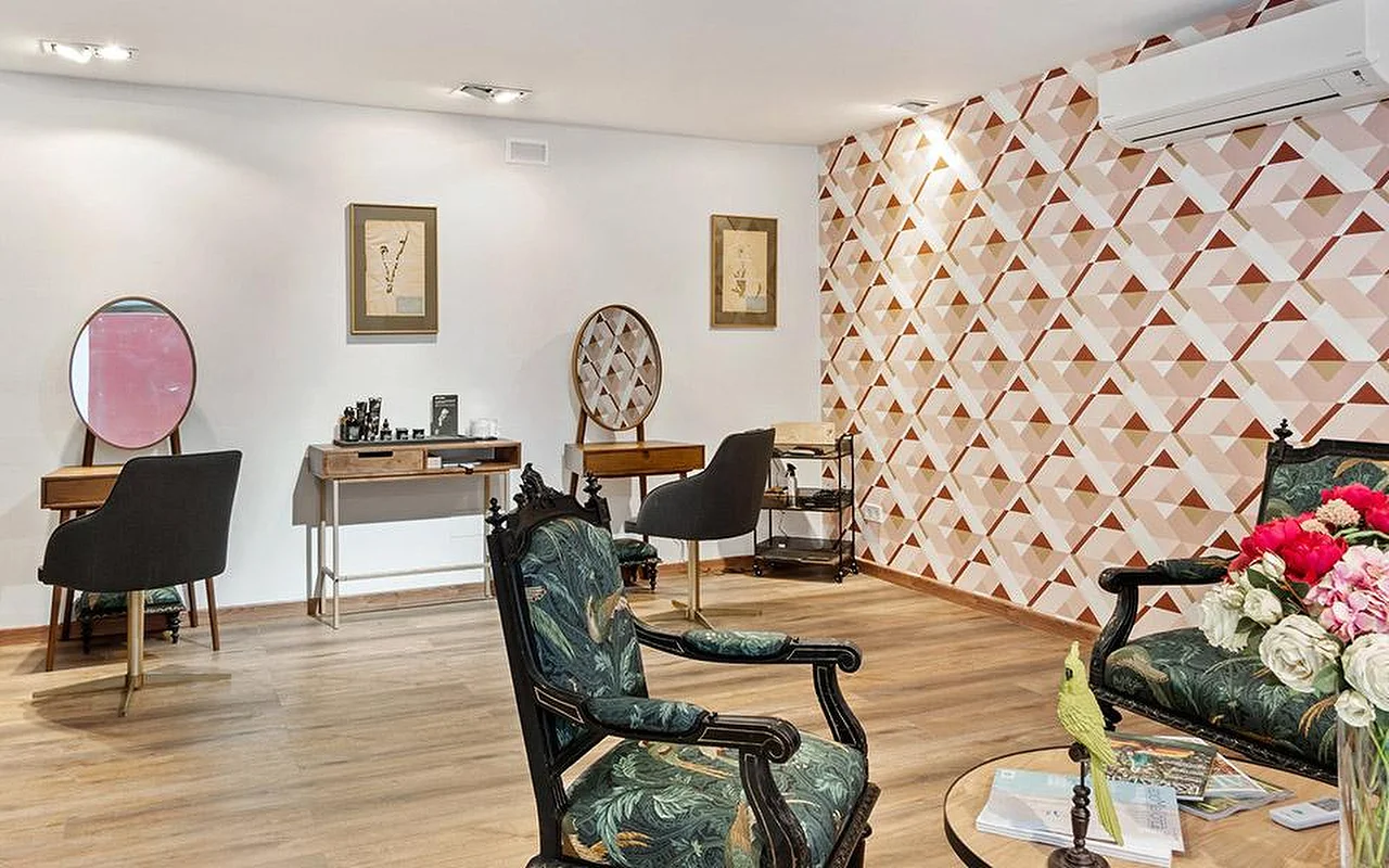 Elegante sala di un salone con una parete decorativa geometrica nei toni del rosa e del terracotta, due poltrone intagliate e rivestite in tessuto botanico verde, postazioni di styling in legno con specchi rotondi e, in primo piano, un vaso di fiori rosa e rossi.