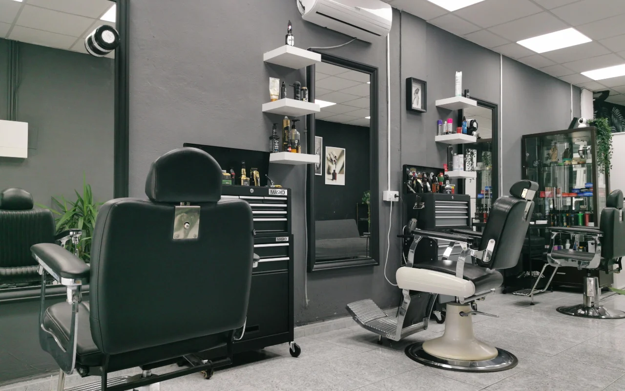 Barbershop tradizionale con pareti grigio antracite, classiche poltrone da barbiere in pelle nera con basi e poggiapiedi cromati, mensole bianche sospese cariche di prodotti per la cura della persona e grandi specchi su un pavimento piastrellato.