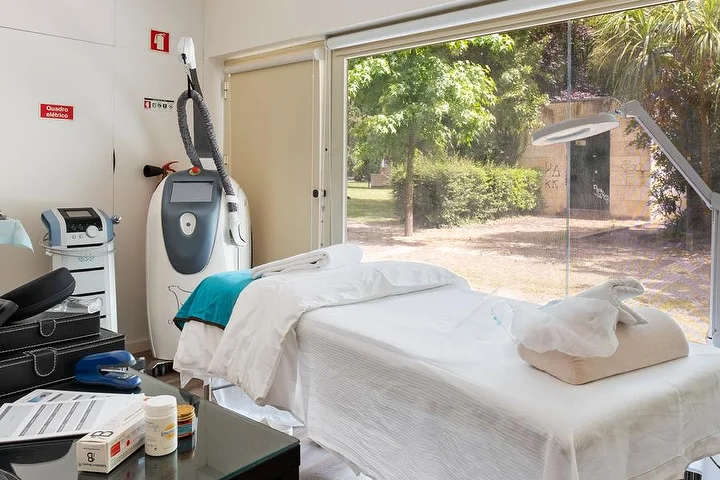 Una cabina per trattamenti con un lettino drappeggiato di bianco accanto a una vetrata a tutta altezza su un giardino verde, un macchinario per trattamenti corpo su piantana, una lampada d'ingrandimento e un tavolino di vetro con prodotti.