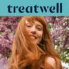 Primo piano ravvicinato di una giovane donna con i capelli rossi e sorridente con la frangetta, con i capelli mossi dal vento davanti a uno sfondo di fiori di lillà rosa. In alto, una fascia azzurra mostra il logo "Treatwell" in blu scuro.