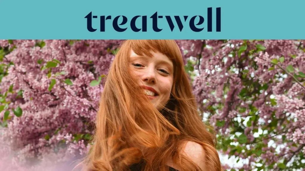 Primo piano ravvicinato di una giovane donna con i capelli rossi e sorridente con la frangetta, con i capelli mossi dal vento davanti a uno sfondo di fiori di lillà rosa. In alto, una fascia azzurra mostra il logo "Treatwell" in blu scuro.