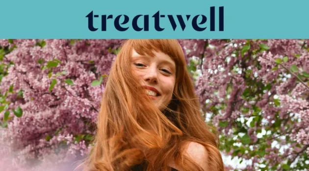 Primo piano ravvicinato di una giovane donna con i capelli rossi e sorridente con la frangetta, con i capelli mossi dal vento davanti a uno sfondo di fiori di lillà rosa. In alto, una fascia azzurra mostra il logo "Treatwell" in blu scuro.