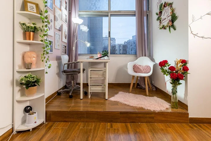 Un piccolo studio di estetica su una pedana di legno rialzata, con una scrivania bianca e una lampada a pinza, una sedia da ufficio e una poltroncina, mensole d'angolo con piante, un vaso di rose rosse e una finestra sulla città.