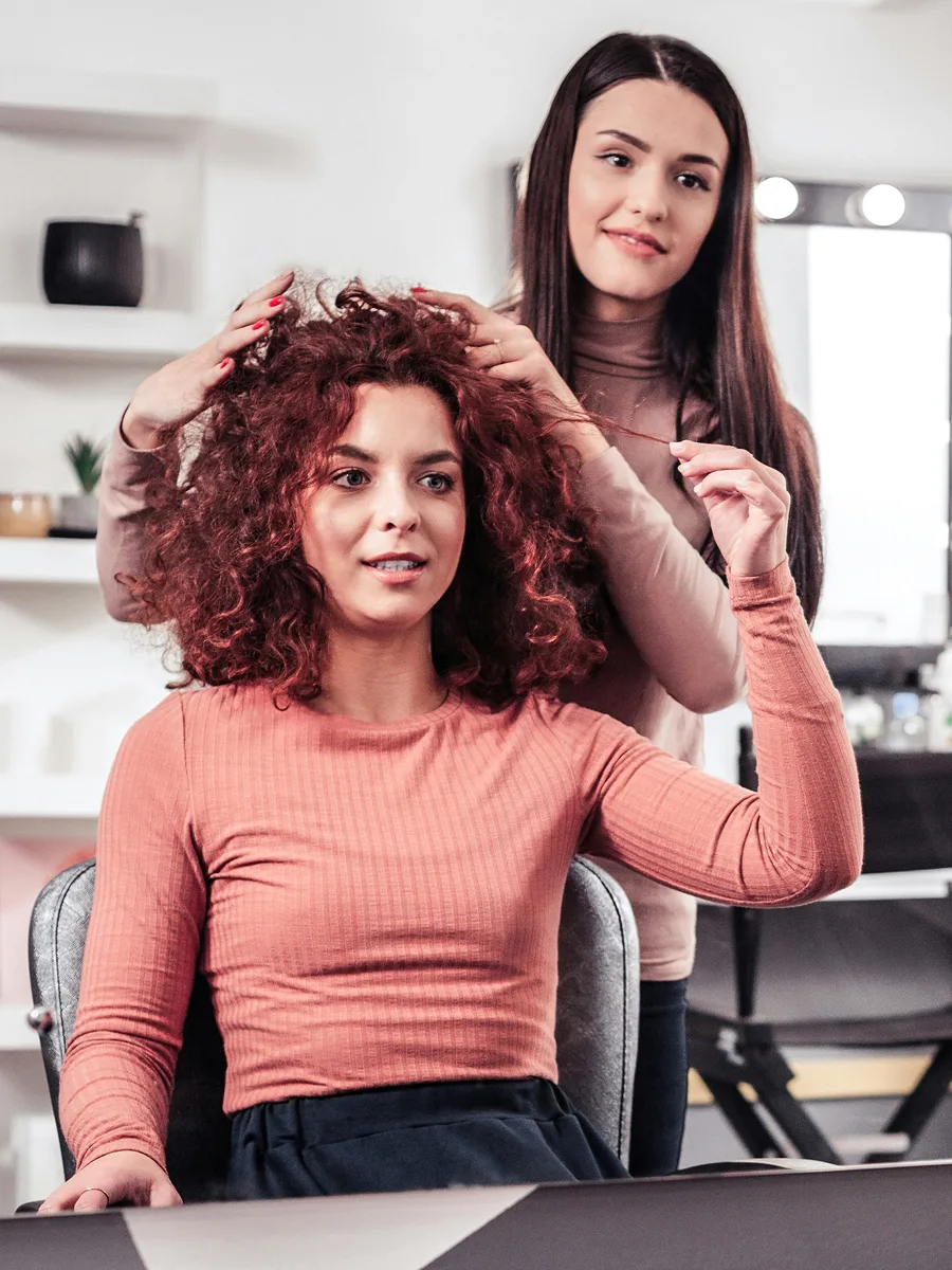 Una cliente con capelli ricci tinti di rosso seduta dal parrucchiere mentre l'hairstylist le illustra il trattamento