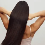 Vista posteriore di una donna con un top bianco a spalline e le braccia alzate mentre passa le dita tra i lunghi capelli castani e lucidi
