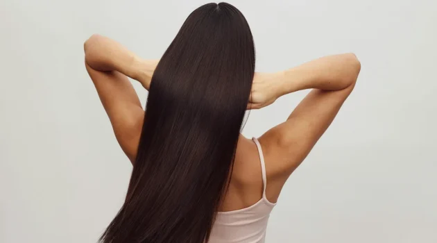 Vista posteriore di una donna con un top bianco a spalline e le braccia alzate mentre passa le dita tra i lunghi capelli castani e lucidi