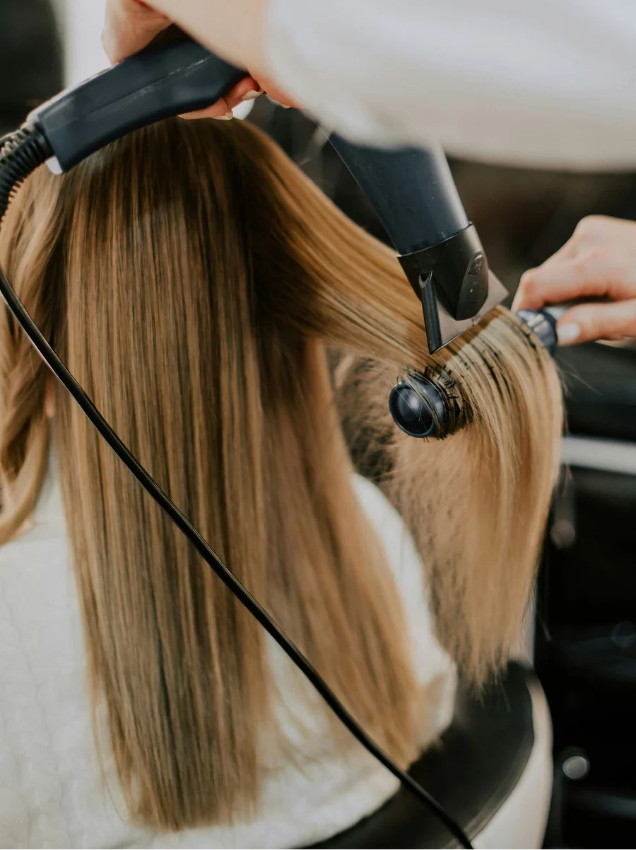 Vista posteriore di una donna dal parrucchiere mentre un hairstylist le asciuga i capelli lisci durante un appuntamento per il trattamento alla cheratina