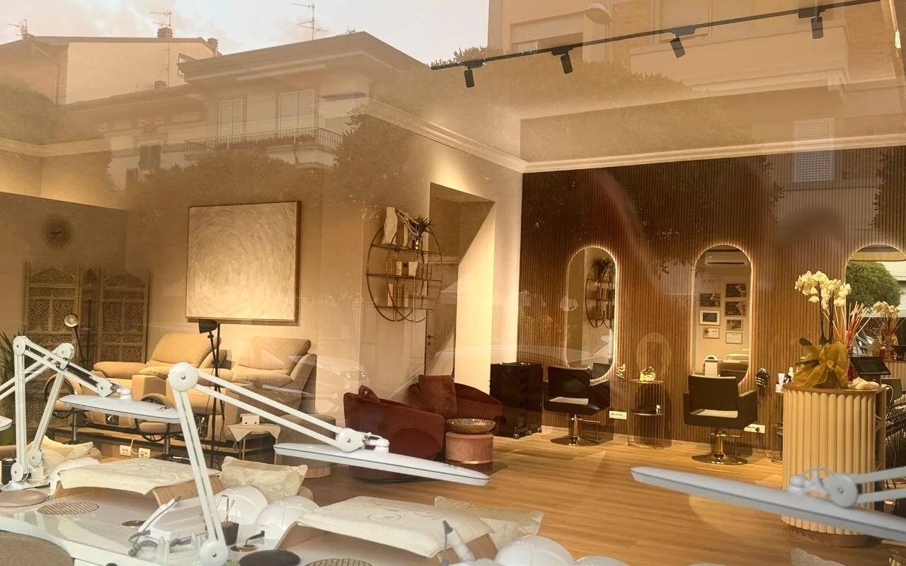 L'interno del salone di Lilian Almanza visto attraverso la vetrina all'ora dorata. Sul lato sinistro, le postazioni per le unghie con lampade regolabili; sulla destra, boiserie in legno caldo, specchi ad arco con cornici retroilluminate e poltrone in velluto.