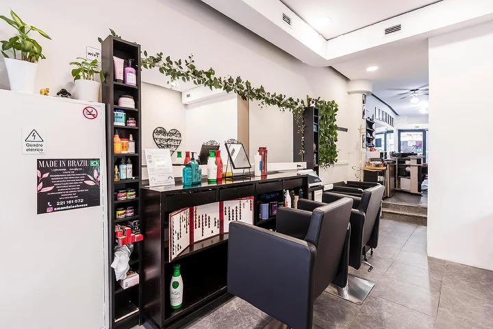 Un salone di capelli stretto e lungo con poltrone nere lungo una parete a specchio, una mensola scura piena di prodotti per capelli, una ghirlanda di verde che ricade sullo specchio e un'insegna Made In Brazil incorniciata su una colonna bianca.