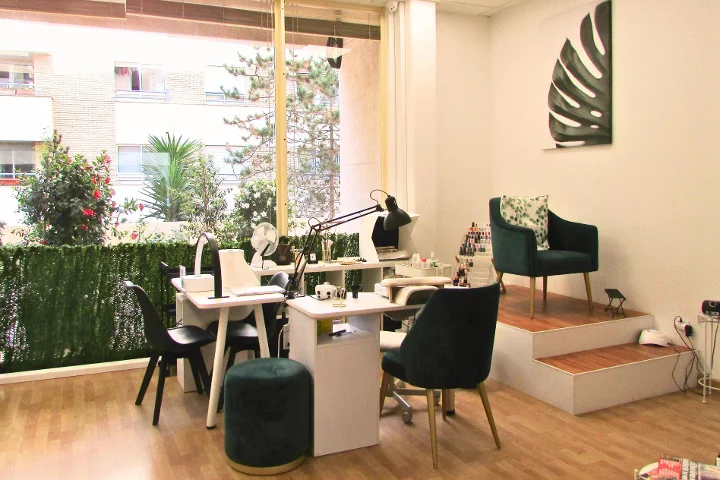 Un'area manicure luminosa con due tavolini bianchi per unghie e relative lampade, poltroncine in velluto verde e uno sgabello abbinato, una tela con foglia di monstera alla parete e ampie finestre su una terrazza con piante.