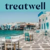 Il lungomare della Piccola Venezia a Mykonos: case imbiancate a calce con balconi di legno colorati a filo dell'acqua, il mar Egeo turchese sullo sfondo e, in primo piano, tavolini da caffè bianchi e verde acqua su una terrazza lastricata in pietra.
