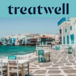 Il lungomare della Piccola Venezia a Mykonos: case imbiancate a calce con balconi di legno colorati a filo dell'acqua, il mar Egeo turchese sullo sfondo e, in primo piano, tavolini da caffè bianchi e verde acqua su una terrazza lastricata in pietra.