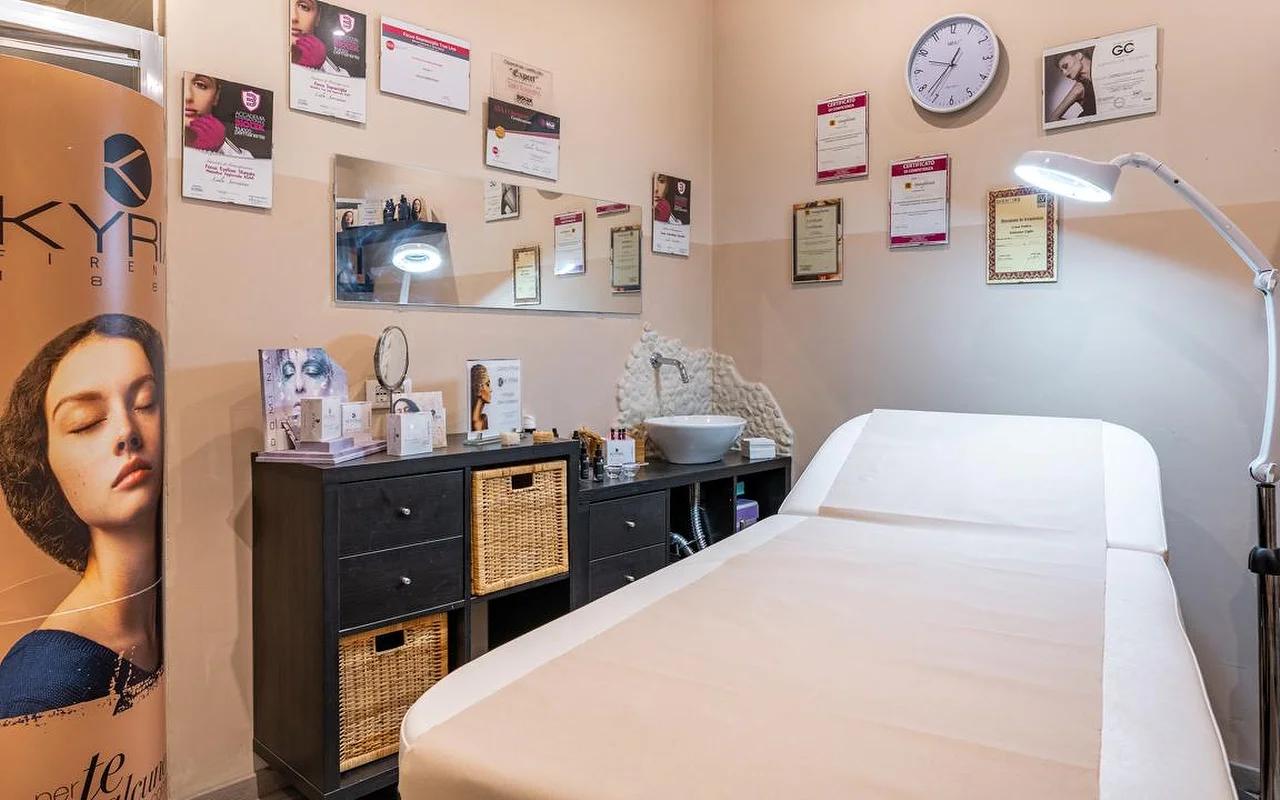 Una cabina trattamenti di Oltre la bellezza con un lettino bianco, una lampada con lente d'ingrandimento regolabile e le pareti rivestite di attestati e diplomi professionali. Sotto a uno specchio ci sono mobili contenitori in vimini scuro, con i prodotti per la pelle disposti sul piano.