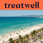 Veduta aerea di Playa de Palma a Maiorca, con una lunga distesa di sabbia dorata, file di ombrelloni, acqua turchese e calma e un lungomare costeggiato di palme che corre lungo la città, con le colline in lontananza sotto un cielo azzurro e limpido.