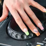 Una mano con l’electric mani appoggiata sul jog wheel di un lettore DJ Pioneer, con french tip al neon in stampa animalier e a spirale