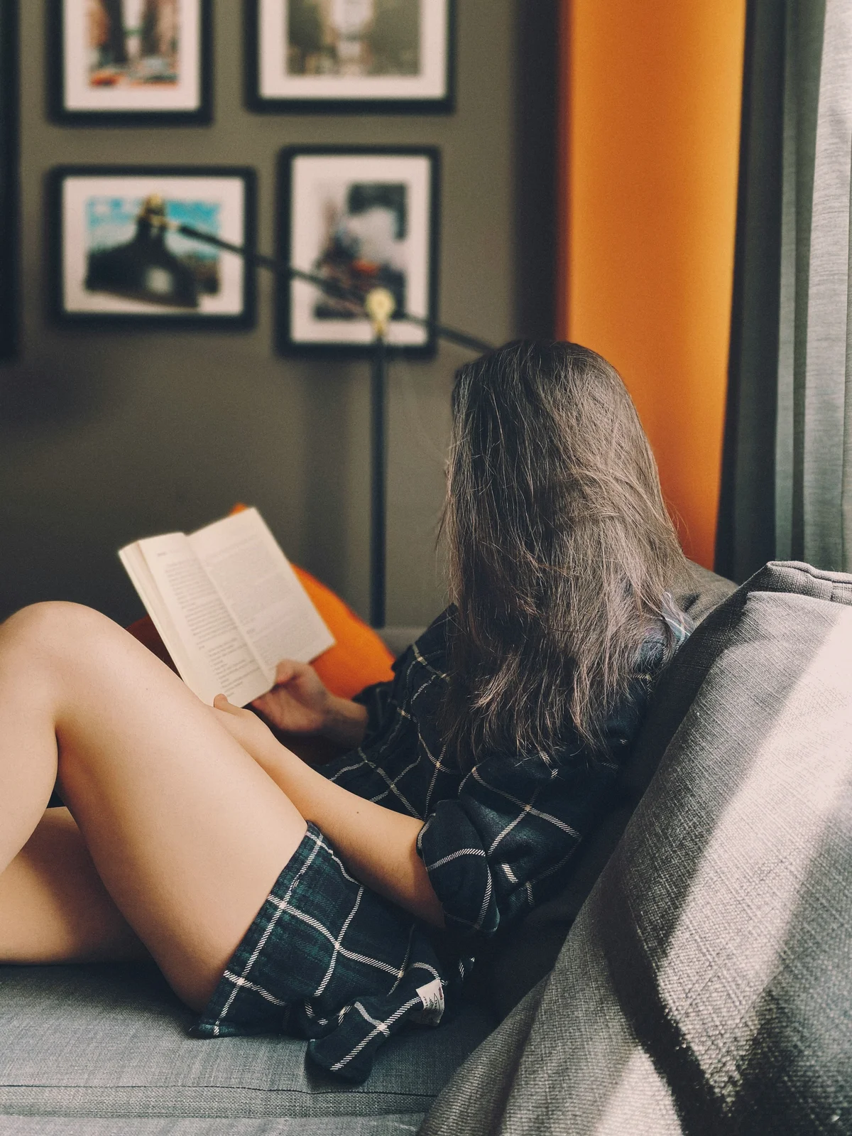 Vista laterale di una donna seduta su un divano che legge un libro, i lunghi capelli le coprono il viso; indossa pantaloncini di cotone larghi e una camicia a quadri verde scuro