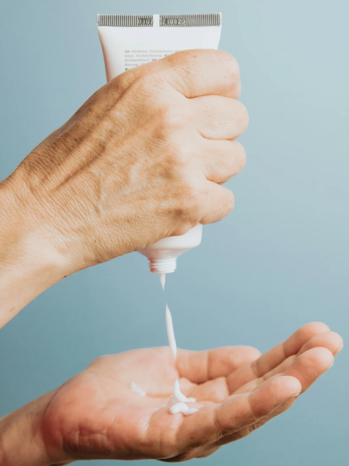 Primo piano di una mano che tiene in verticale un tubetto di crema idratante bianca e ne fa cadere un po' nell'altra mano, sotto
