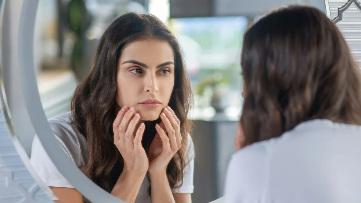 Una donna con lunghi capelli scuri e sopracciglia ordinate e ben definite si guarda allo specchio, con le mani appoggiate delicatamente al viso