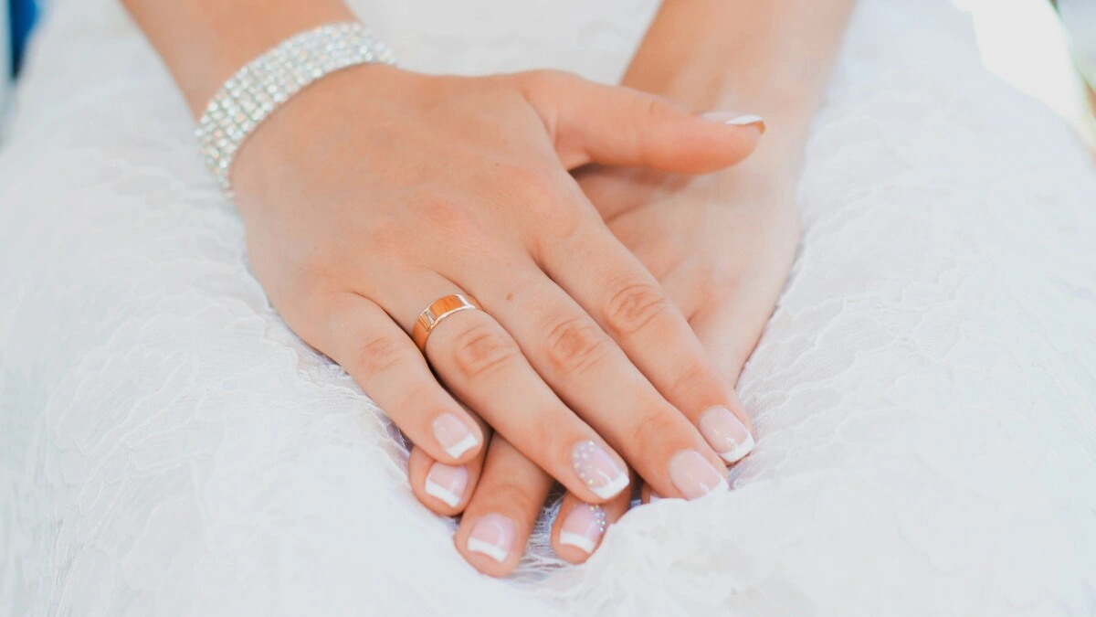 Le mani di una donna riposano in grembo; indossa un abito da sposa in pizzo e una curata French manicure in una combinazione classica di nude e bianco su unghie corte squadrate-ovali, con una delicata decorazione sugli anulari