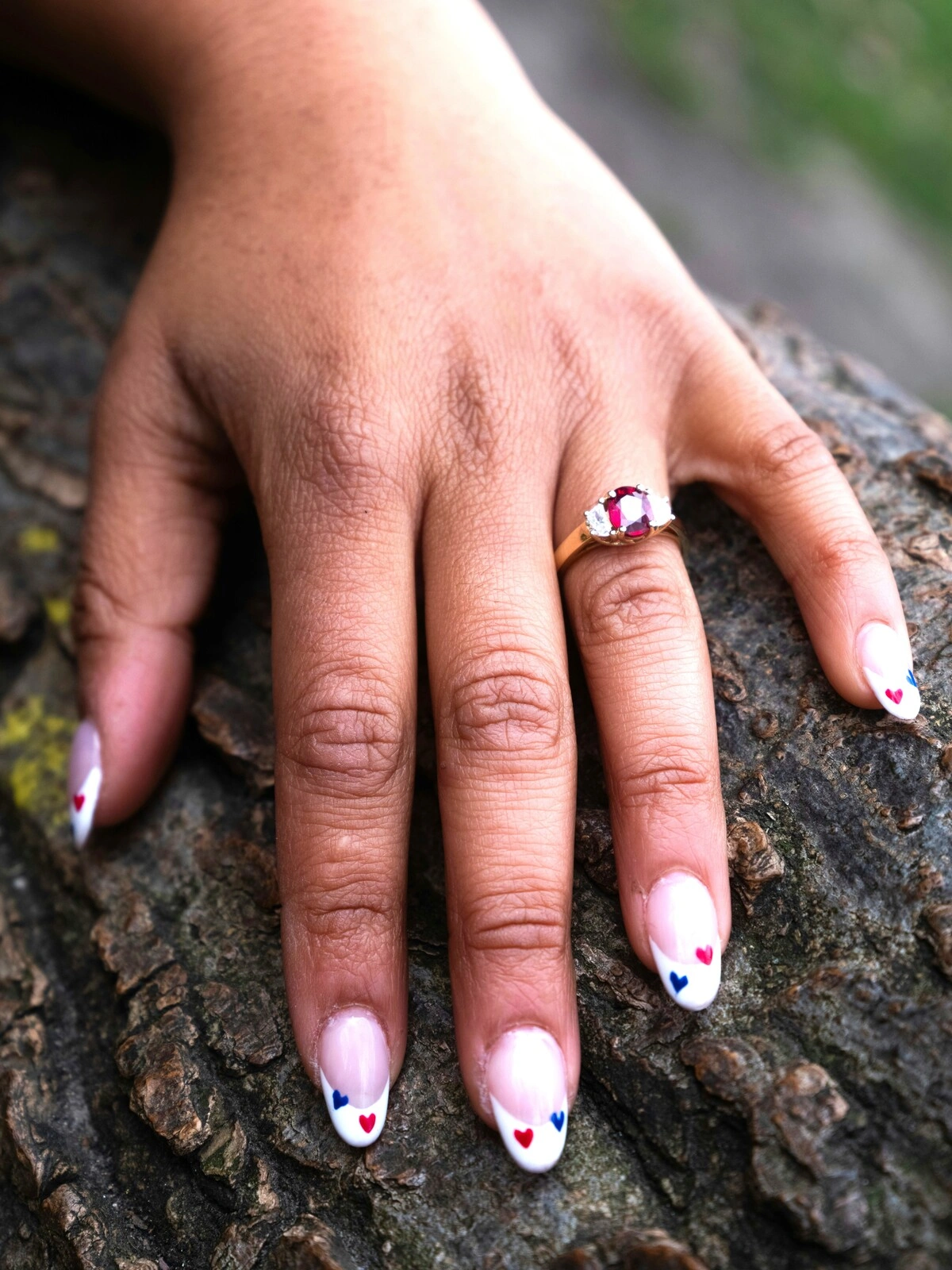 La mano di una donna appoggiata su un ramo mostra una French manicure su unghie ovali, decorate con piccoli cuori rossi e blu