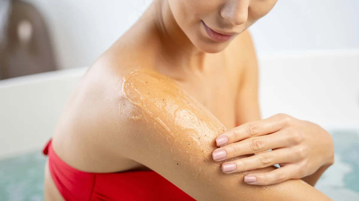 Primo piano di una donna in bikini rosso che applica uno scrub sulla parte alta del braccio in un bagno luminoso e ordinato