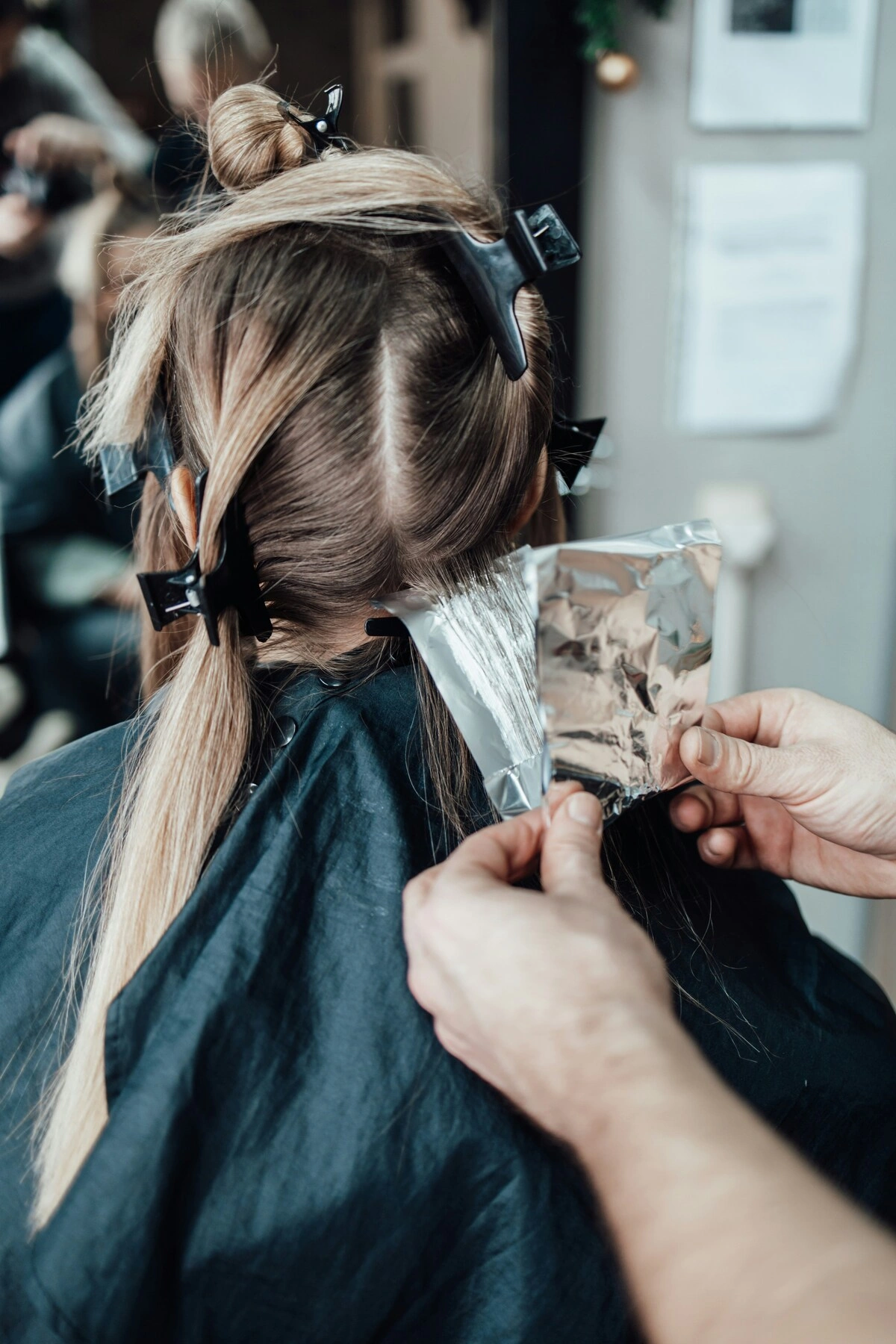 Una cliente vista di spalle su una poltrona da parrucchiere mentre l'hairstylist applica le lamelle tra i capelli per le meches