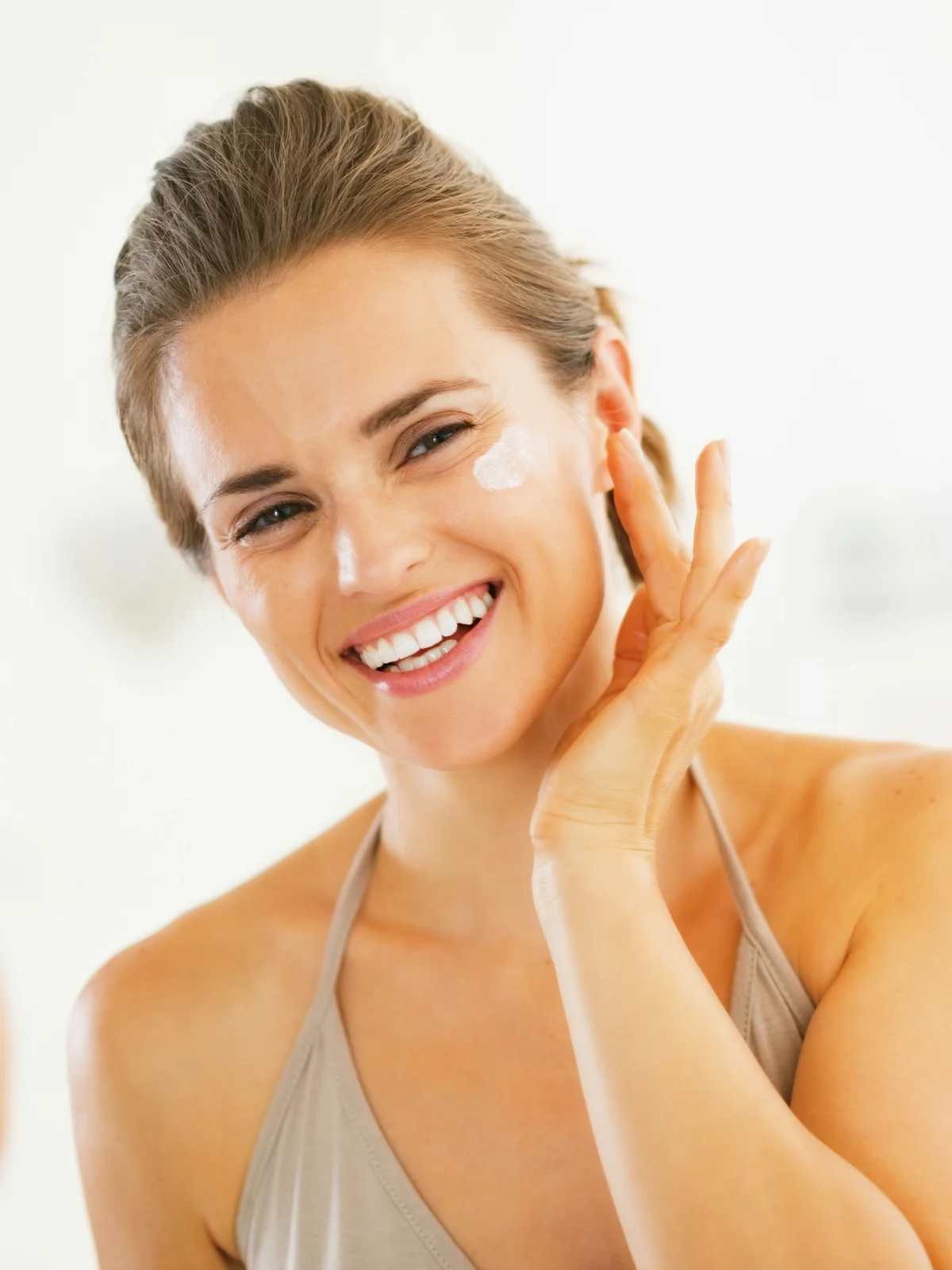 Primo piano di una donna sorridente con i capelli raccolti che applica la protezione solare sul viso