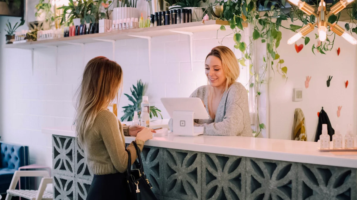 Una donna è al bancone di un salone mentre una receptionist prenota il suo prossimo trattamento; il salone è ordinato, con un bancone bianco, mensole di prodotti ben sistemate e piante e fiori come decorazione