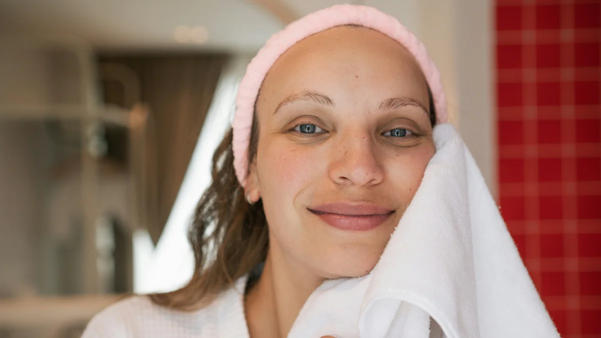Una donna con la pelle pulita e luminosa sorride verso l'obiettivo con una fascia rosa tra i capelli mentre si tampona il viso appena deterso con un asciugamano bianco