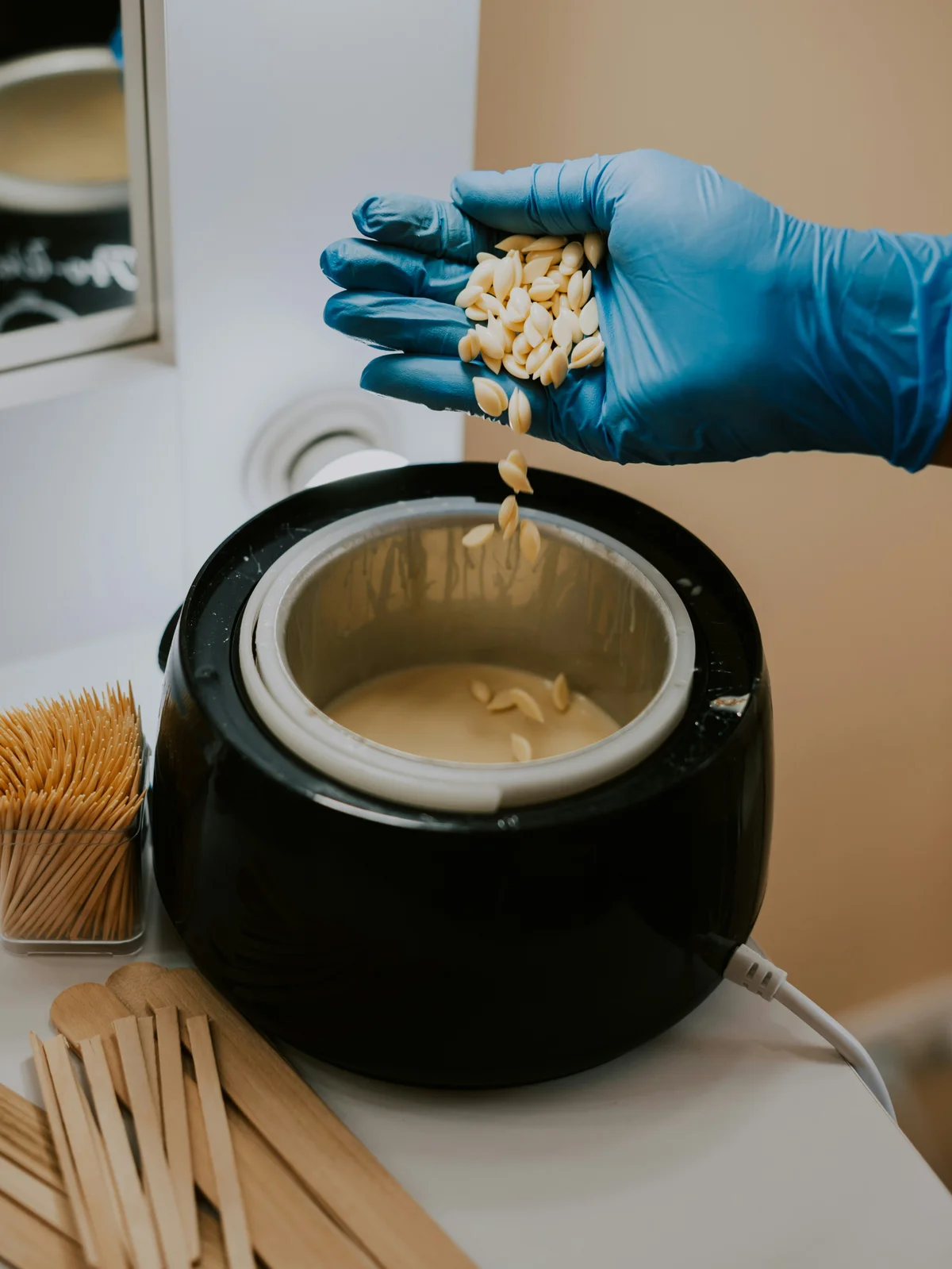 Primo piano della mano guantata di un’estetista che mescola perle di cera calda in un pentolino, pronta per un appuntamento in cabina