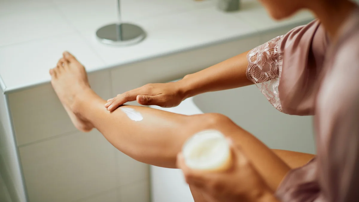 Scatto sopra la spalla di una donna in vestaglia di seta lilla mentre applica una crema idratante sulla gamba appoggiata al piano del bagno