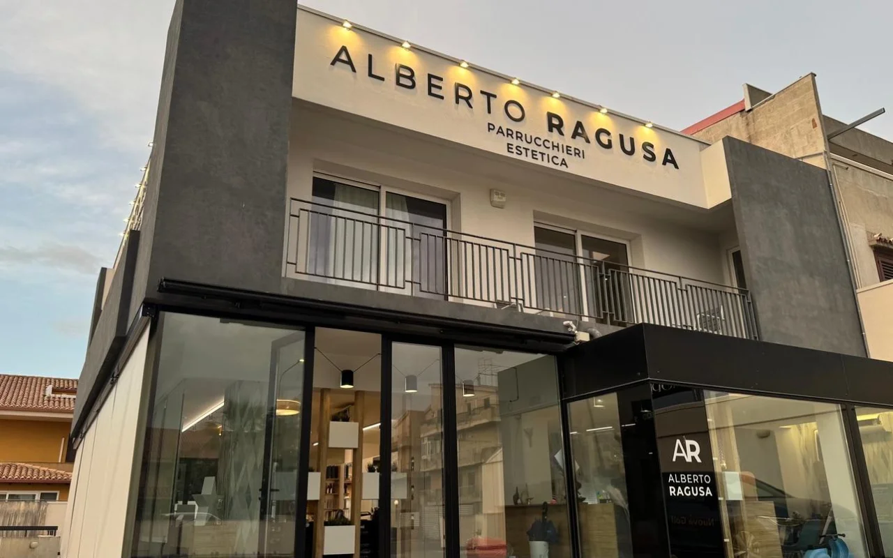 L'esterno di Alberto Ragusa al crepuscolo, un salone su due piani con una grande insegna illuminata che recita 'Alberto Ragusa Parrucchieri Estetica' sopra vetrate dal pavimento al soffitto, con una calda luce che filtra dall'interno.